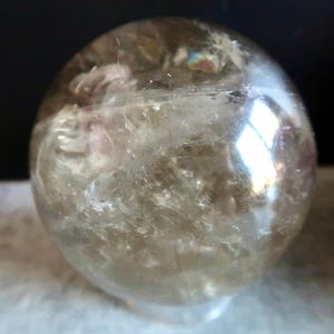 May include: A polished, spherical quartz crystal with a translucent, smoky brown hue. The interior features visible natural inclusions, fractures, and a small, iridescent rainbow-coloured patch reflecting light near the top surface.