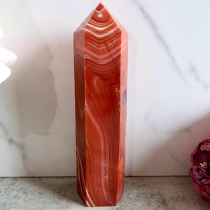 May include: A polished, six-sided crystal tower featuring a vibrant red and orange colour palette with distinct, swirling white banded patterns throughout the stone. The tower has a pointed, faceted top and a smooth, reflective surface finish.