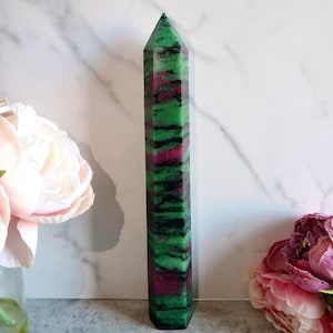 May include: A tall, faceted crystal obelisk with a pointed top. The obelisk features bands of green, black, and red. The crystal is standing upright, with pink and white flowers in the background. The obelisk is a decorative item.