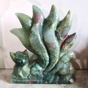 Puede incluir: Una escultura de piedra tallada que representa un zorro con varias colas desplegadas detrás. La figura está hecha de piedra verde moteada con vetas visibles y pequeñas inclusiones rojas, presentando texturas detalladas en forma de hoja.