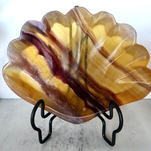 May include: A yellow, purple and brown fluorite shell with a black metal stand. The fluorite is carved into a shell shape and has a smooth, polished surface.