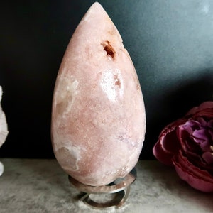 Puede incluir: Un cristal rosa en forma de l&aacute;grima sobre un soporte de metal. El cristal tiene una superficie lisa y pulida con inclusiones blancas y marrones.