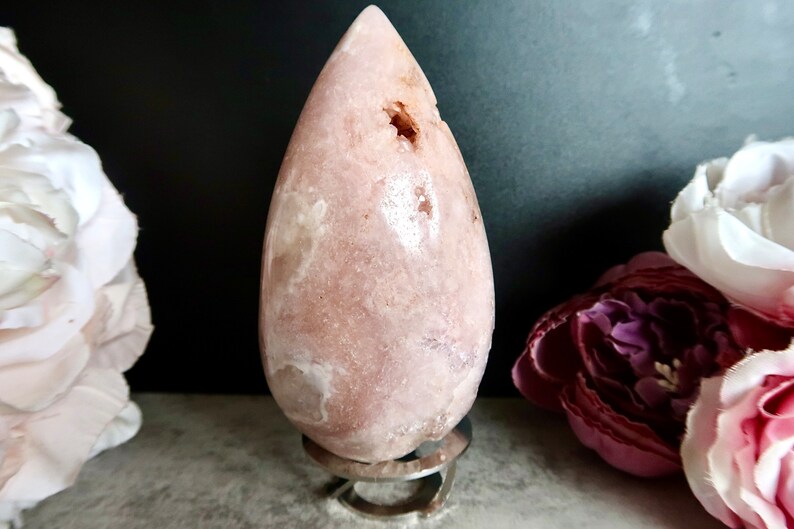 Puede incluir: Un cristal rosa en forma de l&aacute;grima sobre un soporte de metal. El cristal tiene una superficie lisa y pulida con inclusiones blancas y marrones.