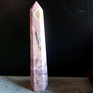 May include: A tall, pink and white crystal obelisk with a pointed top. The stone has a marbled appearance with streaks of grey and tan. The obelisk is standing upright against a dark background.