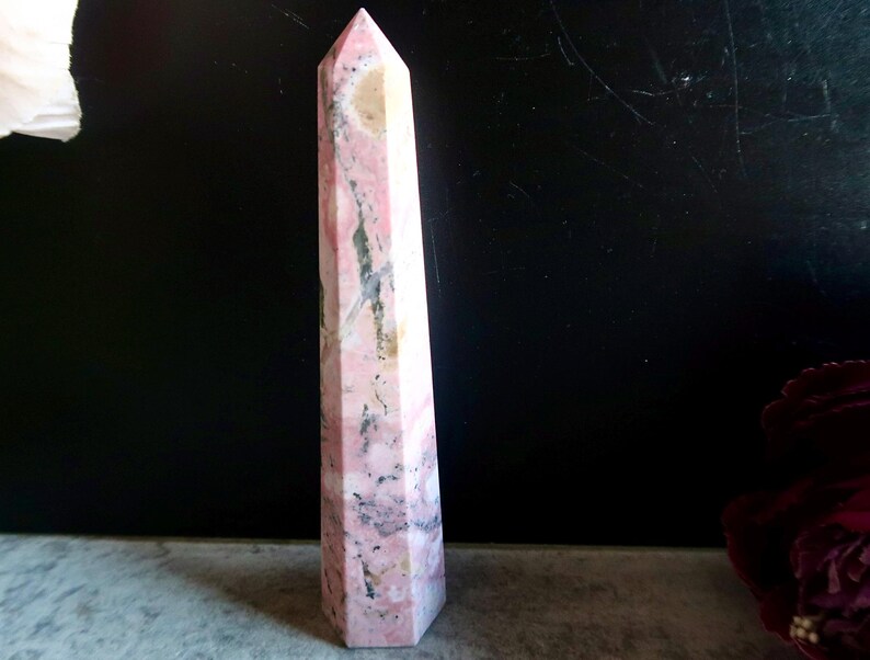 May include: A tall, pink and white crystal obelisk with a pointed top. The stone has a marbled appearance with streaks of grey and tan. The obelisk is standing upright against a dark background.