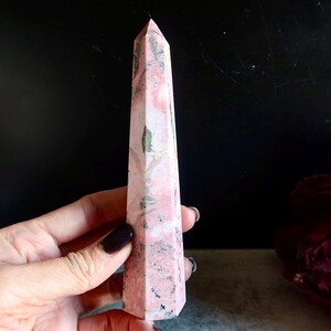 May include: A pink and grey crystal obelisk with a pointed top. The crystal has a marbled appearance with varying shades of pink and grey. The obelisk is held in a hand, with a dark background and a purple flower in the background.