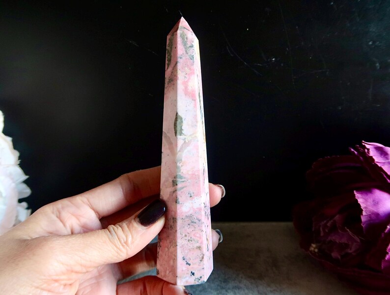 May include: A pink and grey crystal obelisk with a pointed top. The crystal has a marbled appearance with varying shades of pink and grey. The obelisk is held in a hand, with a dark background and a purple flower in the background.