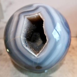 May include: A polished spherical agate stone featuring concentric bands of light grey, white, and dark blue. The centre of the sphere contains a deep, irregular cavity lined with small, sparkling, translucent crystals that create a textured contrast.