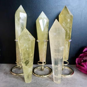 Puede incluir: Se muestran cinco puntas de cristal de cuarzo amarillo-verdoso de varios tamaños. Cada cristal está montado sobre un soporte de metal dorado. Los cristales tienen un aspecto translúcido y están dispuestos sobre un fondo oscuro con una flor rosa.