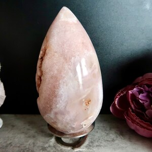 Puede incluir: Una geoda de &aacute;gata rosa y blanca tallada en forma de l&aacute;grima, expuesta en un soporte de metal. La geoda tiene una superficie lisa y pulida y un interior natural de tipo cristal.