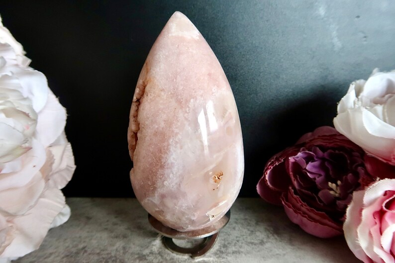 Puede incluir: Una geoda de &aacute;gata rosa y blanca tallada en forma de l&aacute;grima, expuesta en un soporte de metal. La geoda tiene una superficie lisa y pulida y un interior natural de tipo cristal.
