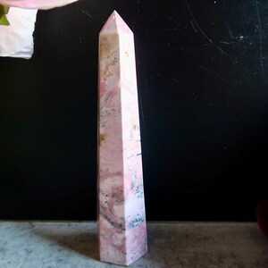 May include: A pink and white obelisk crystal, with a pointed top. The crystal has a marbled effect with darker pink and grey streaks. The background is dark, with a hint of a flower in the upper left corner.