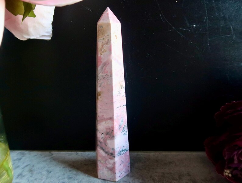 May include: A pink and white obelisk crystal, with a pointed top. The crystal has a marbled effect with darker pink and grey streaks. The background is dark, with a hint of a flower in the upper left corner.