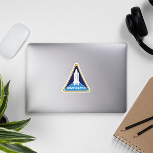 May include: A triangular sticker featuring a white space shuttle graphic centered on a blue background with a gold border. The text SPACE SHUTTLE is printed in white capital letters at the bottom of the triangle, which is applied to the center of a laptop.