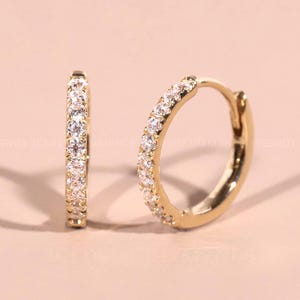 May include: A pair of gold hoop earrings, each encrusted with small, clear gemstones. The earrings are displayed on a light pink surface, with a shadow cast to the side. The hoops are a classic jewelry design.