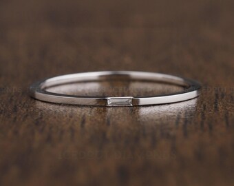 May include: A thin silver band ring with a small, rectangular, clear diamond set in the center.
