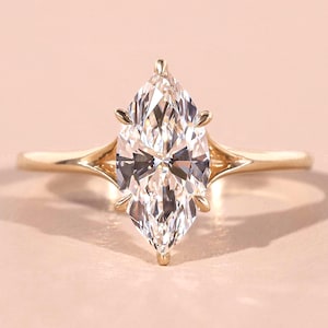 Könnte beinhalten: Ein Verlobungsring mit einem Diamanten im Marquiseschliff und einem goldenen Band. Der Diamant ist mit vier Krappen gefasst, und das Band hat ein geteiltes Schaftdesign. Der Ring wird auf einer hellrosa Oberfläche präsentiert.