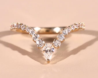 May include: A gold-colored ring with a V-shaped design. The ring is adorned with numerous small, clear gemstones, with a larger pear-shaped gemstone at the center of the V. The ring is set against a soft, neutral background.