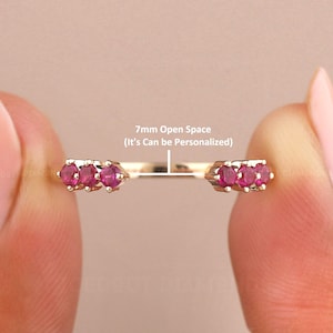 May include: A gold-colored ring with three small, round, red gemstones on each end. The ring has a 7mm open space that can be personalized. The ring is held between two fingers.