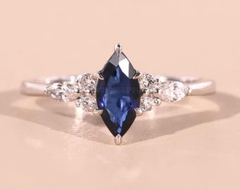 Op de afbeelding: Een zilveren ring met een opvallende, marquise-geslepen, diepblauwe edelsteen in het midden, geflankeerd door kleinere, heldere edelstenen. Het ontwerp van de ring is elegant en modern, tegen een zachte, neutrale achtergrond.