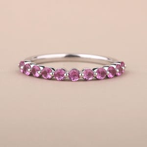 Könnte beinhalten: Ein silberner Ring mit einer Reihe kleiner, runder, rosa Edelsteine. Die Edelsteine sind gleichmäßig um das Band angeordnet. Der Ring ist vor einem neutralen Hintergrund platziert.