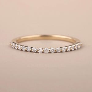 May include: A gold band ring featuring small, round, clear gemstones. The ring is presented against a neutral backdrop, highlighting its understated elegance. The gemstones are uniformly spaced around the ring's circumference.