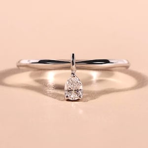 Op de afbeelding: Een delicate zilveren ring met een peervormige diamant hanger. De ring heeft een dunne, gepolijste band, met de diamant die aan een kleine verticale staaf hangt. De diamant schittert tegen een neutrale achtergrond.