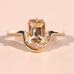 Puede incluir: Un engaste de anillo dorado con un centro de talla esmeralda y un diseño curvo único. El anillo se encuentra sobre un fondo rosa suave, resaltando sus intrincados detalles y su artesanía. El diseño del anillo es moderno y elegante.