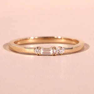 Puede incluir: Un anillo de banda dorado con tres pequeños diamantes. El diamante central es rectangular, flanqueado por dos diamantes redondos. El anillo se presenta sobre un fondo rosa suave, resaltando su delicado diseño.