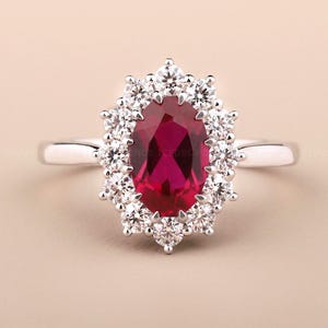 Könnte beinhalten: Ein silberner Ring mit einem ovalen, tiefroten Edelstein, umgeben von einem Kranz aus kleineren, klaren Steinen. Das Design des Rings ist klassisch und elegant, wobei der Schwerpunkt auf der satten Farbe des zentralen Edelsteins und dem Glanz der umliegenden Steine liegt.
