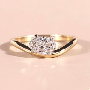 May include: A gold ring featuring a large, oval-cut diamond. The band has a distinctive, curved shape. The diamond is clear and brilliant, held by four prongs. The ring is presented on a pale pink surface.