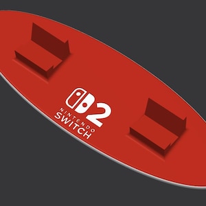 May include: A red Nintendo Switch dock with a white border. The dock has two raised platforms and the Nintendo Switch logo and the number 2 in white. The dock is designed to hold the Nintendo Switch console.