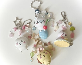 May include: A collection of keychains featuring cartoon characters. The keychains have white, yellow, and pink figures with beaded accents, stars, and charms. One keychain has a white character holding a cherry. Another has a yellow character with a brown mane.
