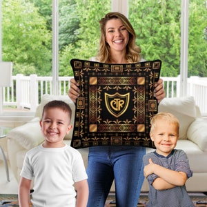May include: A woman holds a black and gold decorative pillow with a gold shield and the letters "CTR" inside. Two children stand on either side of her. The pillow has a geometric pattern.