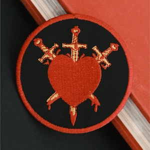 May include: A black and red embroidered patch with three gold swords piercing a red heart. The patch is circular and has a black border.