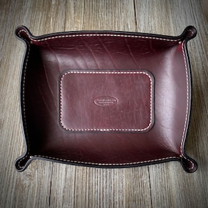 May include: A burgundy leather valet tray with white stitching and a small leather patch with the text "Russell Leather Goods Handmade" in white.