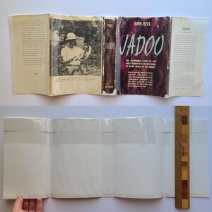 May include: An open book titled "Jadoo" by John Keel, with a dark red cover and the text "The Astounding Story of One Man's Search into the Mysteries of Black Magic in the Orient." The book is open to reveal interior pages with text and a black and white photograph. A wooden ruler is visible.