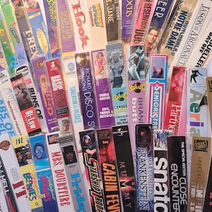 May include: A collection of VHS tapes with various titles and colorful labels. Titles include "Godzilla," "Ferris Bueller's Day Off," "Mrs. Doubtfire," and fitness videos. The tapes are arranged in a fan-like pattern, showcasing a variety of movie genres.
