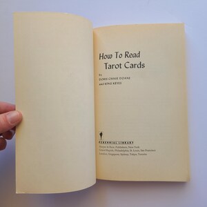 Puede incluir: Libro abierto titulado "How To Read Tarot Cards" de Doris Chase Doane y King Keyes. El libro tiene una cubierta y p&aacute;ginas de color crema. Una mano sostiene el libro abierto.