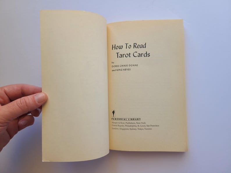 Puede incluir: Libro abierto titulado "How To Read Tarot Cards" de Doris Chase Doane y King Keyes. El libro tiene una cubierta y p&aacute;ginas de color crema. Una mano sostiene el libro abierto.