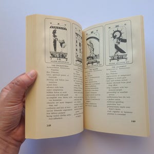 Puede incluir: Un libro abierto que muestra ilustraciones de cartas del tarot y texto. Las p&aacute;ginas est&aacute;n abiertas por una mano, revelando tres cartas con dibujos detallados y descripciones. Las p&aacute;ginas del libro son de color crema y el texto es negro.
