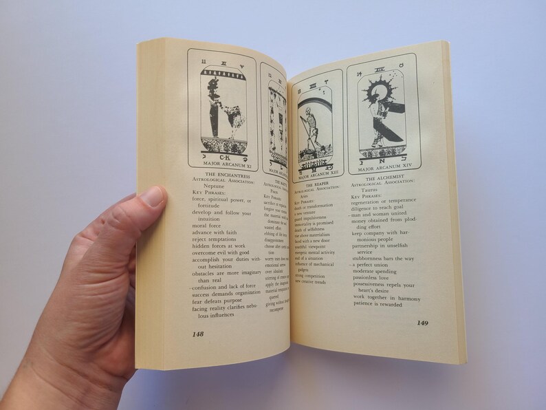 Puede incluir: Un libro abierto que muestra ilustraciones de cartas del tarot y texto. Las p&aacute;ginas est&aacute;n abiertas por una mano, revelando tres cartas con dibujos detallados y descripciones. Las p&aacute;ginas del libro son de color crema y el texto es negro.
