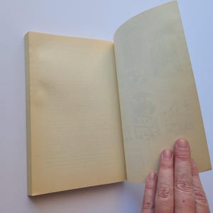 Puede incluir: Un libro abierto con p&aacute;ginas amarillentas. El lado izquierdo est&aacute; cerrado, el derecho abierto, revelando ilustraciones tenues. Una mano sostiene las p&aacute;ginas abiertas.