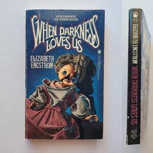 May include: A vintage paperback book titled "When Darkness Loves Us" by Elizabeth Engstrom. The cover features a distressed doll illustration in a red dress against a dark blue background. The spine displays the title and author.