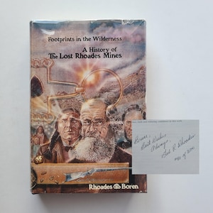 May include: A book titled "Footprints in the Wilderness: A History of The Lost Rhoades Mines" with illustrations of historical figures and mining equipment. Includes a handwritten note that reads "Best Wishes Always, Hal R. Rhoades #80 of 300."