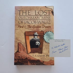 May include: A book titled "THE LOST DUTCHMAN MINE OF JACOB WALTZ Part 1: The Golden Dream" with a vintage map design. The cover features an illustration of a man holding a skull. A handwritten note is also present.