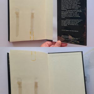 Pu&ograve; includere: Un libro aperto con copertina nera e pagine bianco sporco. La pagina di destra ha del testo, mentre quella di sinistra presenta delle macchie. La sovraccoperta del libro &egrave; visibile, con testo sul contenuto e il design del libro.