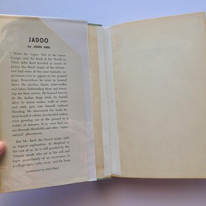 May include: An open book titled "Jadoo" by John Keel. The book's cover is visible, with text and a clear protective cover. The pages are off-white, and the book is held open, revealing the text on the inside.