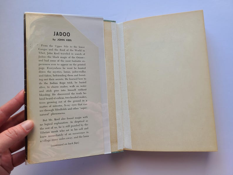 May include: An open book titled "Jadoo" by John Keel. The book's cover is visible, with text and a clear protective cover. The pages are off-white, and the book is held open, revealing the text on the inside.