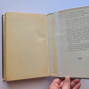 May include: An open book with aged, yellowed pages. The right page has printed text in black, with a small illustration at the bottom. The left page is blank. The book is held open by a hand.